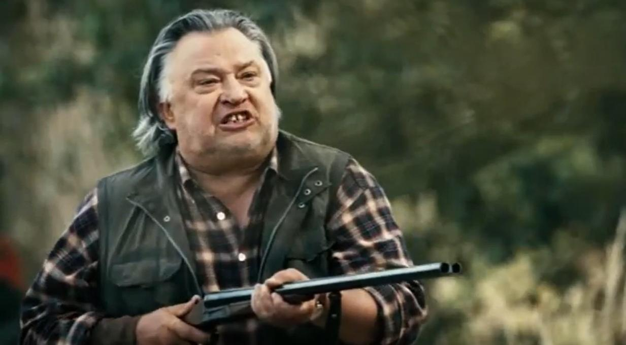 Ein Mann mit grauem Haar und kariertem Hemd hält ein Gewehr.