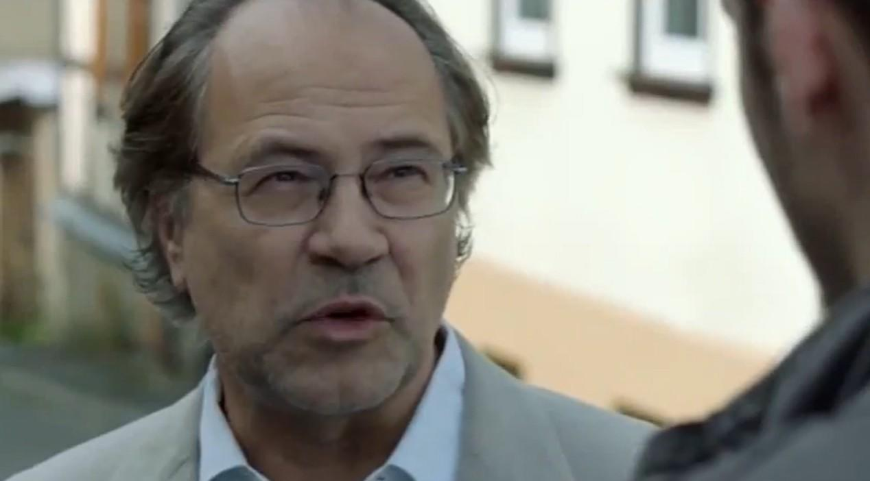 Älterer Mann mit Brille spricht. Im Hintergrund unscharfes Gebäude.