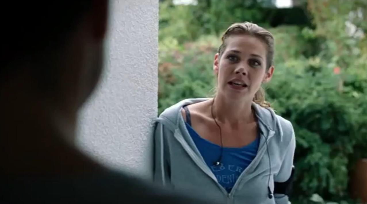 Eine Frau mit braunen Haaren in einem blauen Top und grauer Kapuzenjacke spricht.
