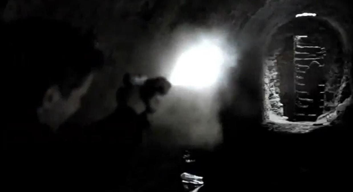 Zwei Personen beleuchten einen dunklen Tunnel mit einer Taschenlampe, der zu Steintreppen führt.