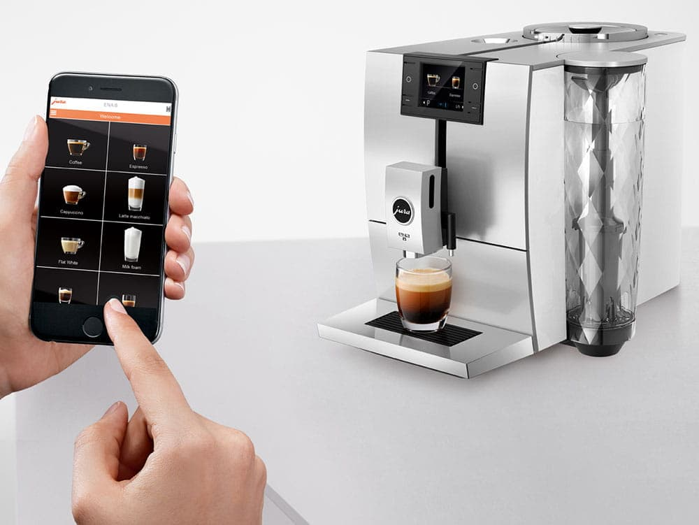 Ręka trzyma smartfon z aplikacją kawową obok srebrnej maszyny do kawy ze szklanką espresso.