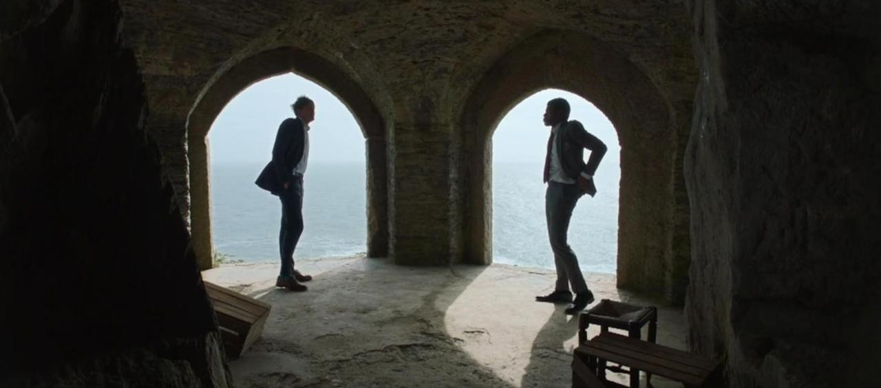 Zwei Männer in Anzügen stehen in einem steinernen Torbogen mit Blick auf das Meer.