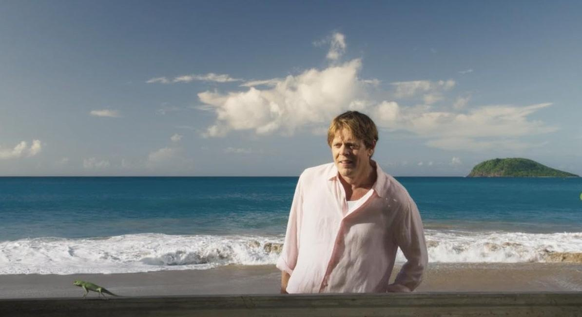 Ein Mann steht am Strand, eine Echse läuft vorbei. Ein kleines Inselchen im Meer.