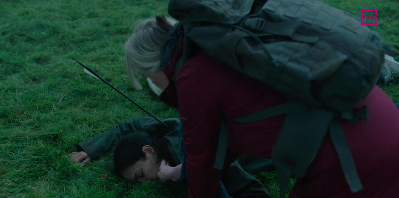 Eine Frau liegt im Gras, ein Pfeil ragt aus ihrem Rücken. Eine andere Frau mit Rucksack kniet über ihr.