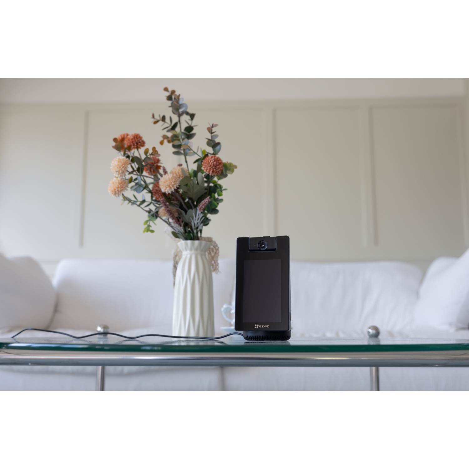 A black smart display speaker sits on a glass table in front of a white sofa.