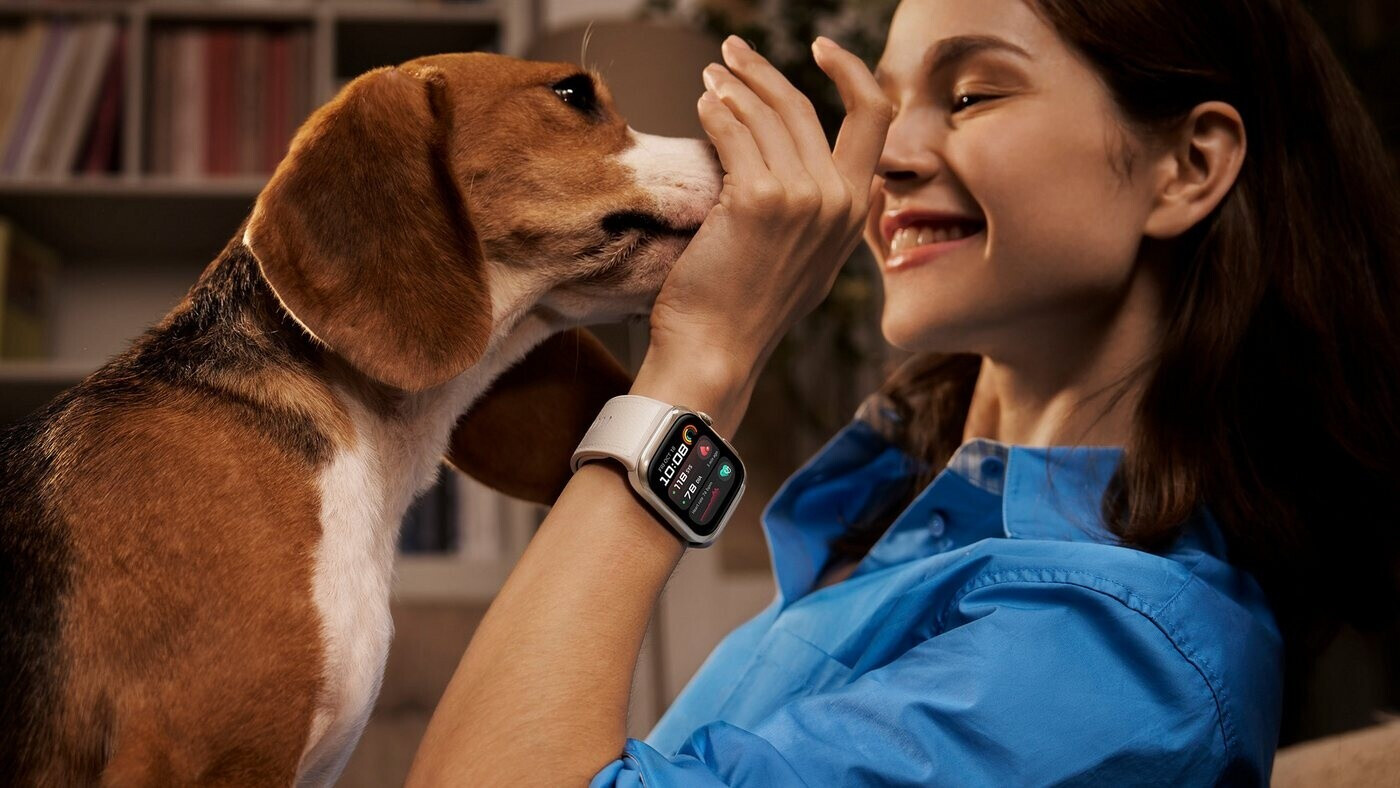 Egy beagle kutya nyalogatja egy okosórát viselő mosolygó nő arcát.