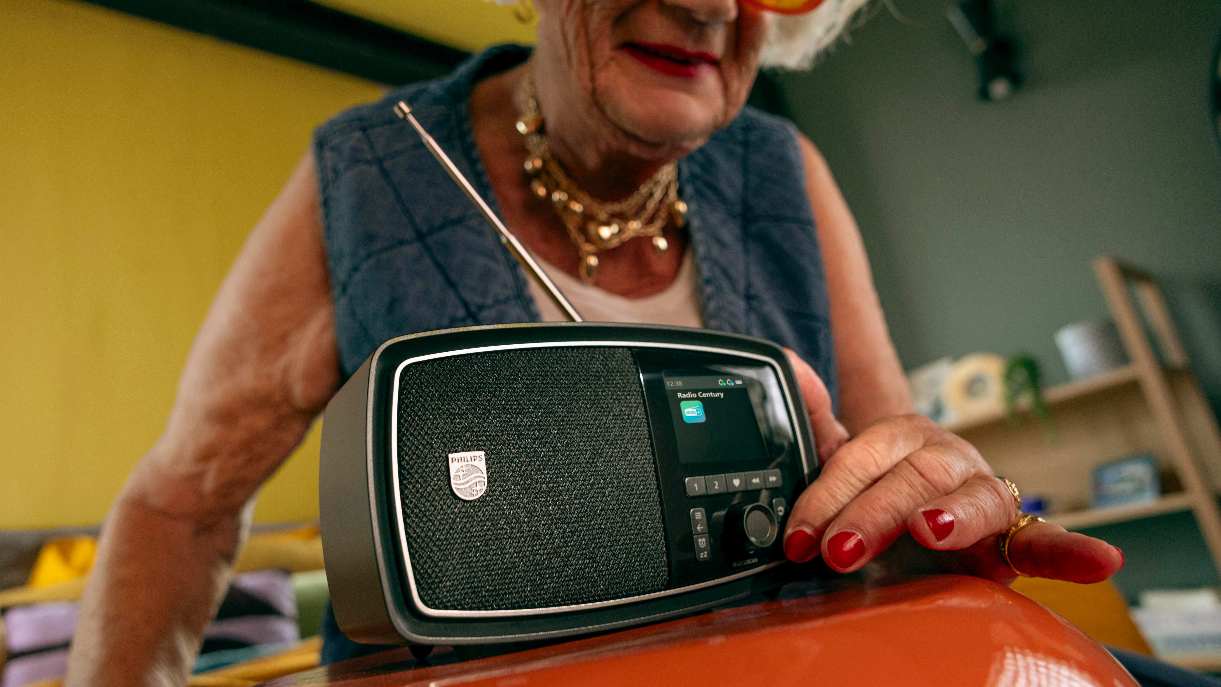 Ältere Frau mit Schmuck stellt ein Radio ein, das auf einem orangefarbenen Tisch steht.