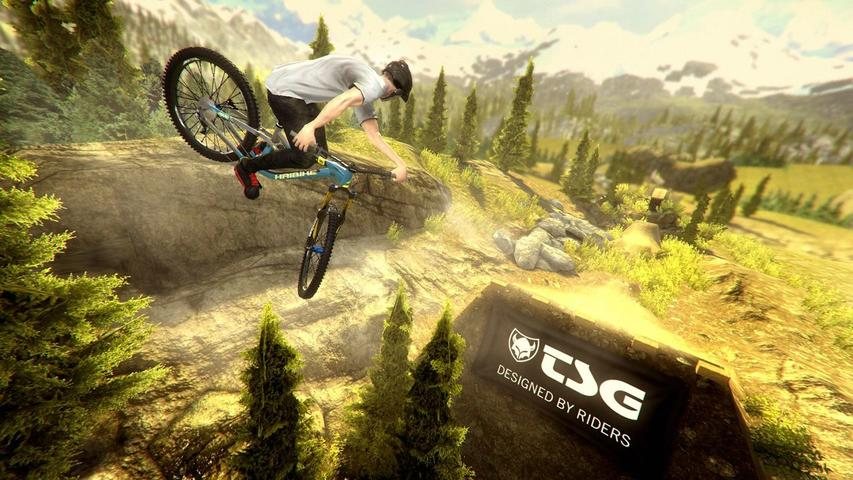 Mountainbiker in der Luft bei Sprung mit Bergkulisse und Bäumen.