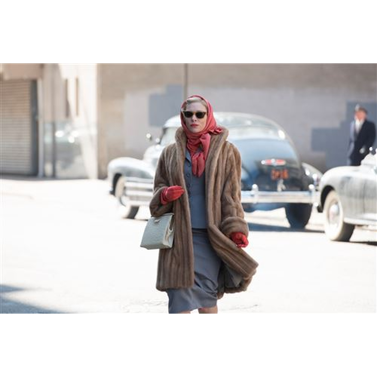 Woman in fur coat, headscarf and sunglasses, holding a purse, in front of cars.