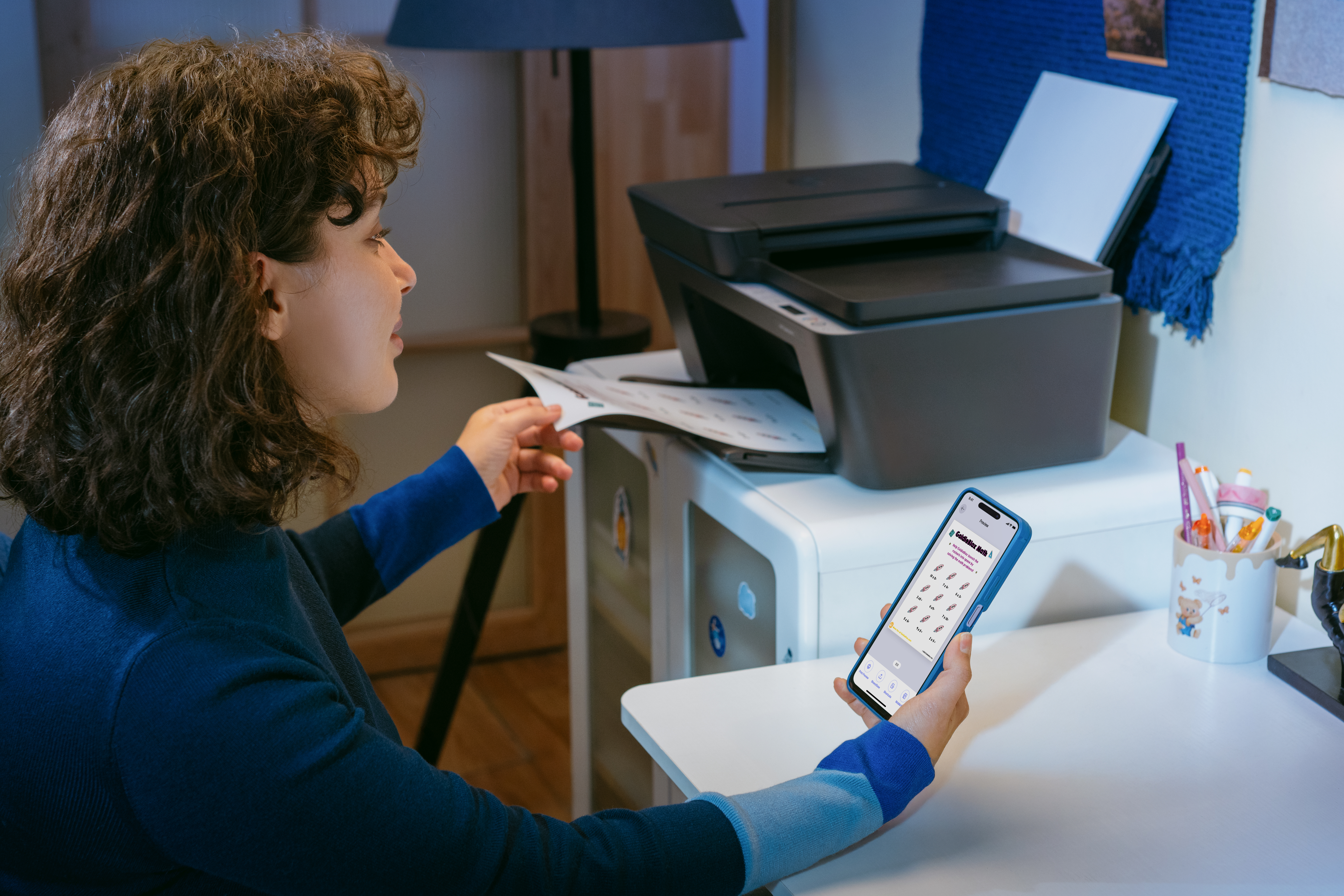 Kobieta z kręconymi włosami używa drukarki i smartfona. Drukarka jest biało-szara.