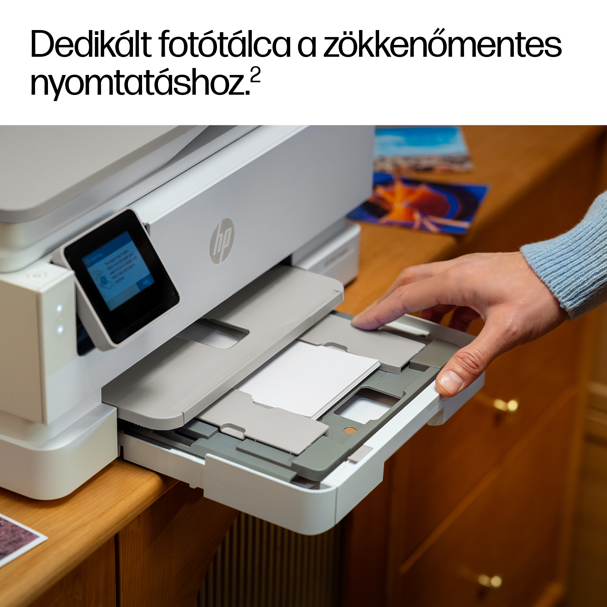 Egy kéz húz ki egy papírkazettát egy fehér nyomtatóról, fényképek az asztalon.