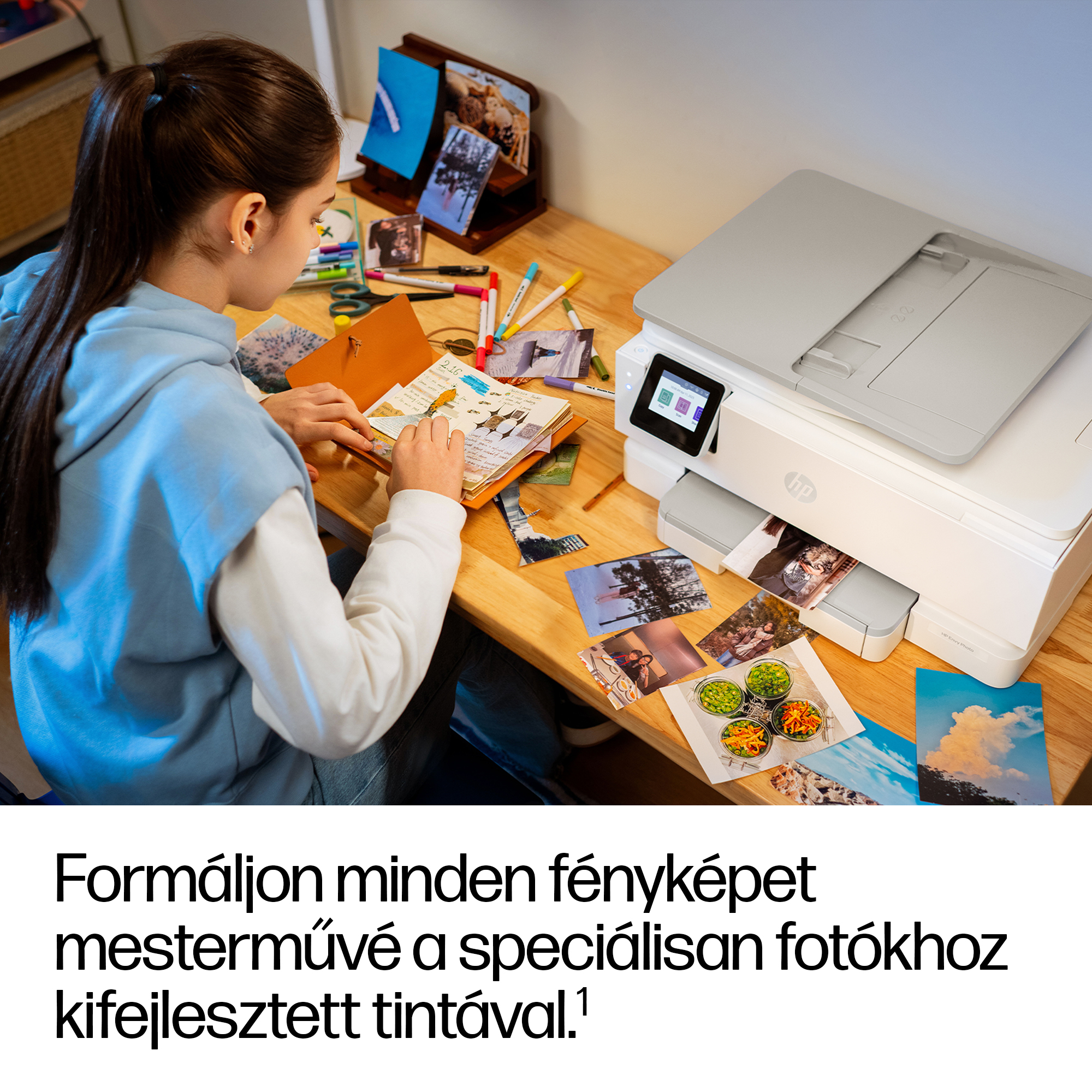 Egy fiatal nő fotókkal és tollakkal díszít egy jegyzetfüzetet egy nyomtató mellett.