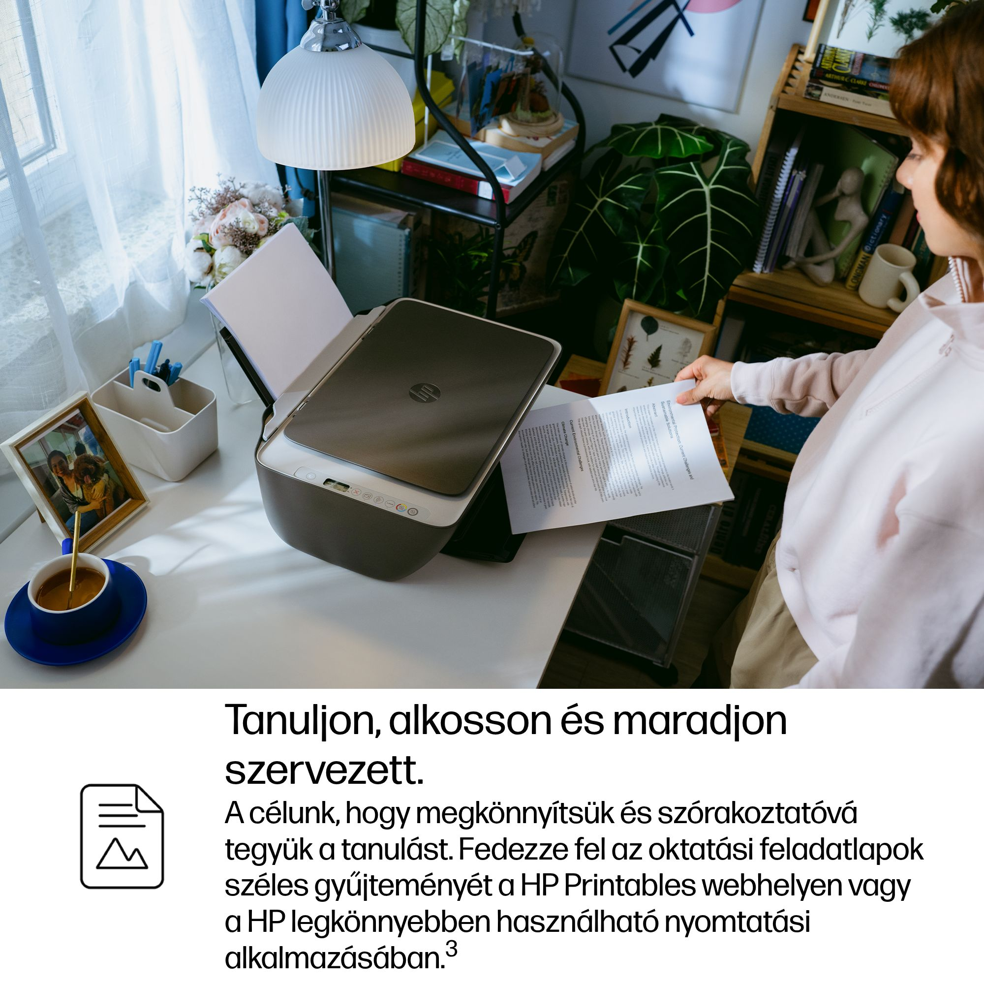 Egy személy dokumentumot tart egy nyomtató, bögre és képkeret mellett.