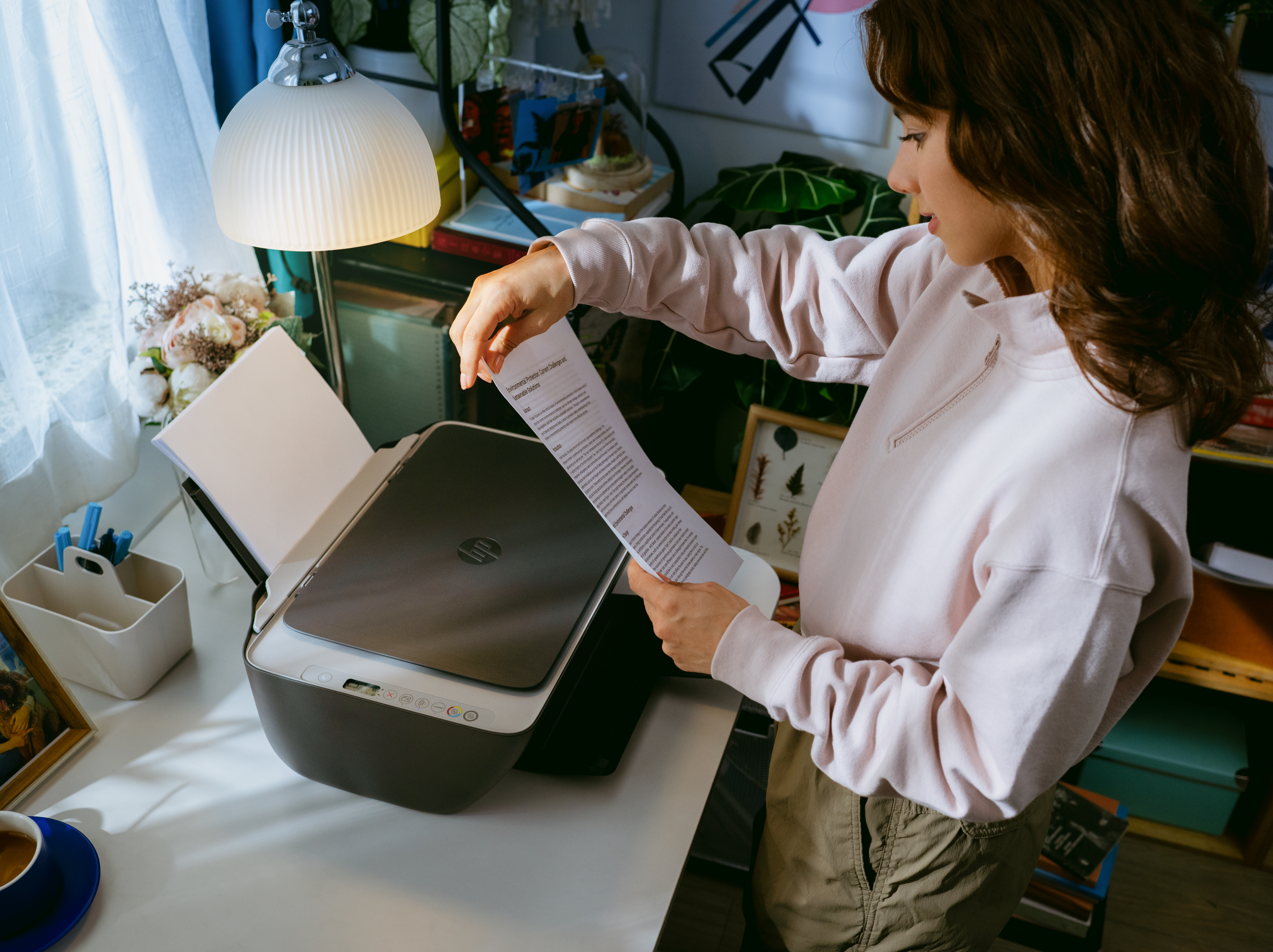 Frau im rosa Sweatshirt legt Papier in Drucker auf Schreibtisch mit Lampe und Kaffee.