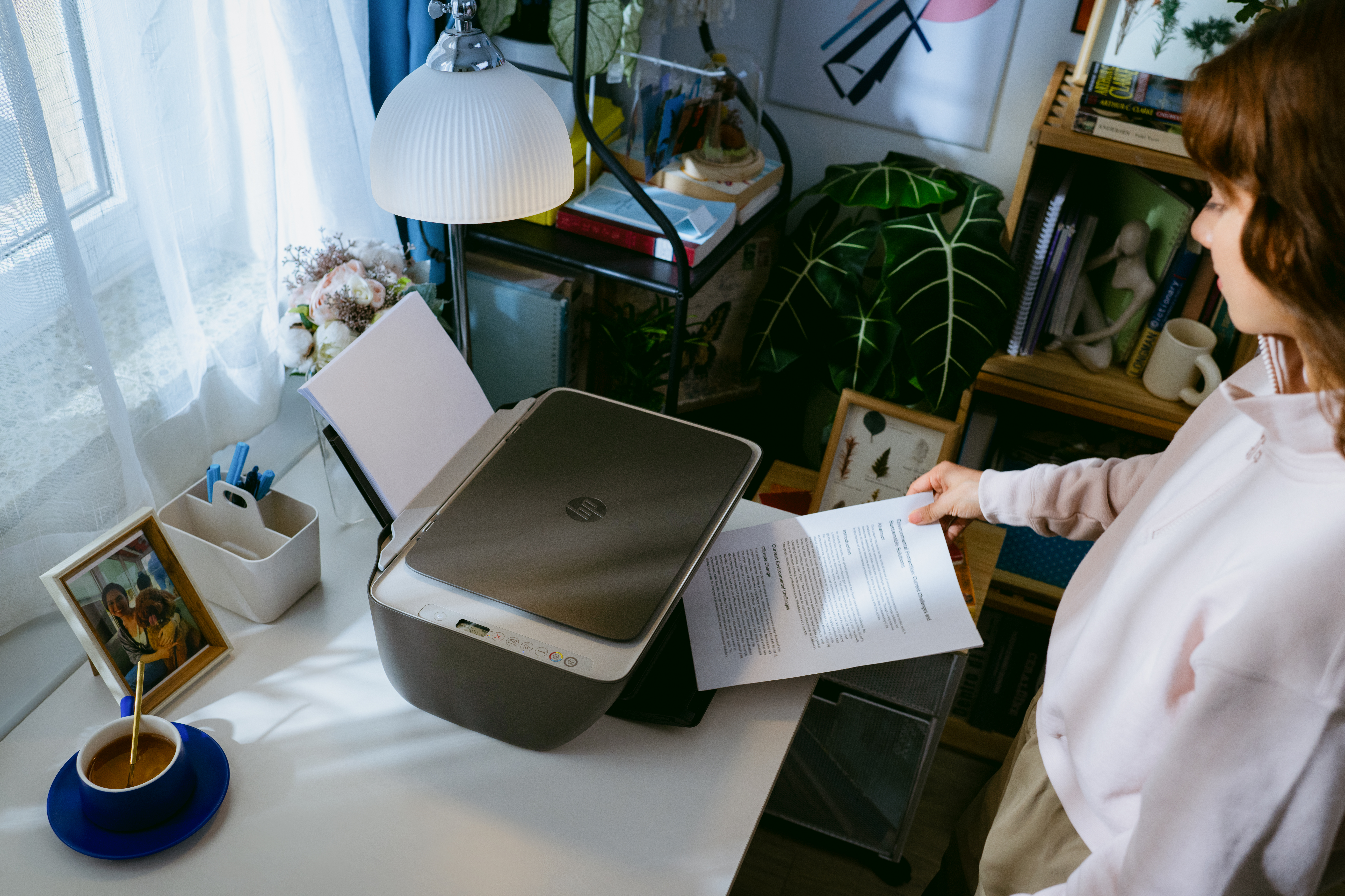 Eine Frau legt ein Blatt Papier in einen Drucker. Auf dem Schreibtisch stehen eine Kaffeetasse und ein Bilderrahmen.