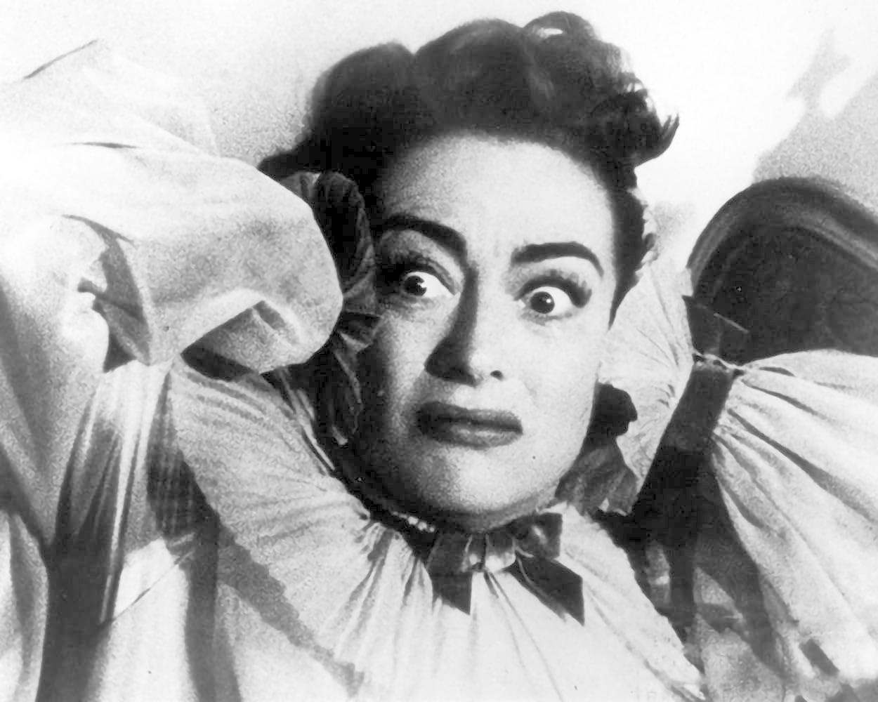 Eine Frau mit weit aufgerissenen Augen und geschocktem Ausdruck, trägt ein Rüschenkleid.