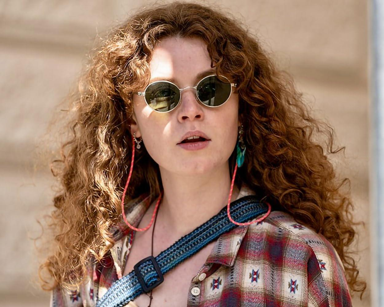 Frau mit roten Locken, Sonnenbrille, kariertem Hemd und auffälligem Ohrring.