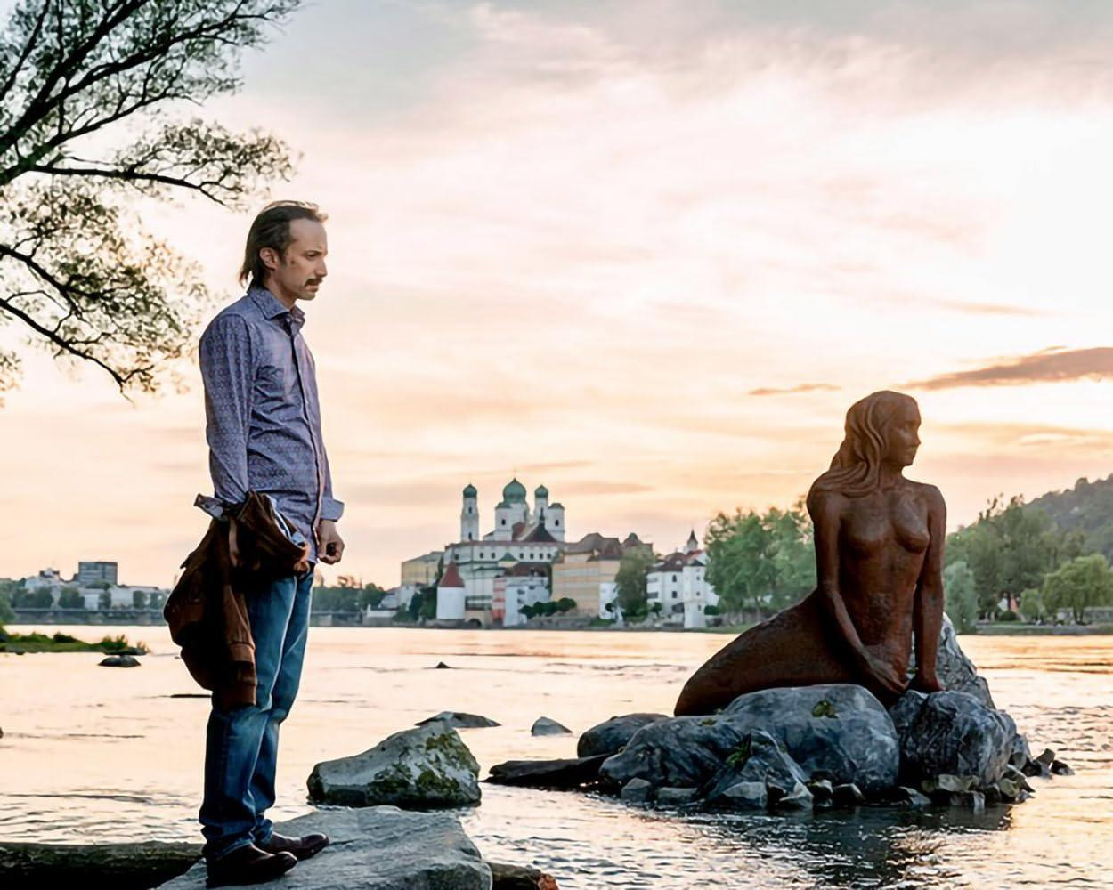 Mann steht neben Meerjungfrauenstatue am Flussufer bei Sonnenuntergang.