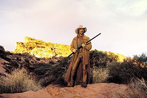 Ein Mann im Western-Look mit Hut und Gewehr steht in felsiger Landschaft.