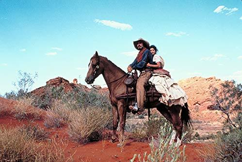 Mann und Frau reiten auf einem Pferd in einer Wüstenlandschaft mit roten Felsen.