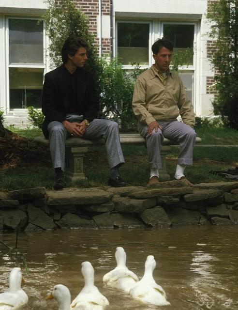 Zwei Männer sitzen auf einer Bank vor einem Gebäude. Enten schwimmen in einem Teich.