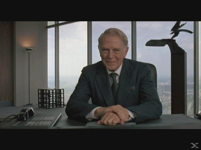 Älterer Mann im Anzug sitzt am Schreibtisch vor einem Fenster mit Stadtblick.