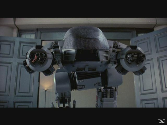 Ein grauer Roboter mit Waffen an den Armen steht vor einer Tür.