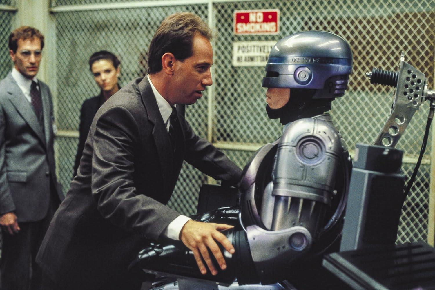 Ein Mann im Anzug spricht mit RoboCop. Im Hintergrund sind zwei Personen.