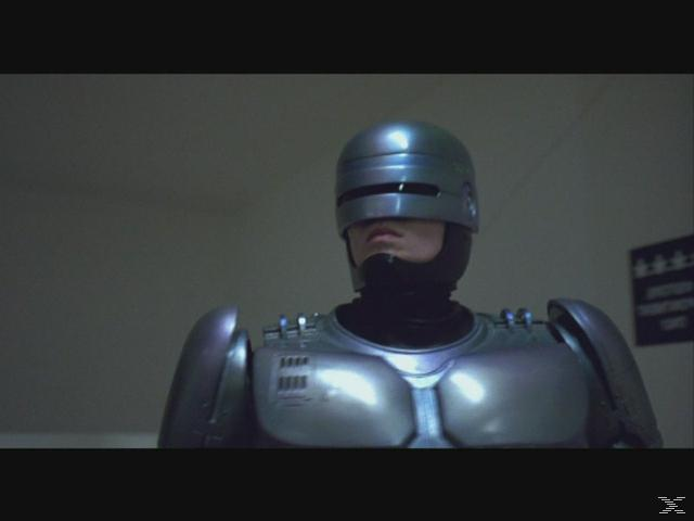 RoboCop in silberner Rüstung steht vor einer grauen Wand.