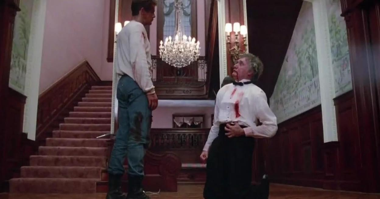 Zwei Männer in blutbefleckter Kleidung in einem opulenten Flur mit Treppen und Kronleuchter.