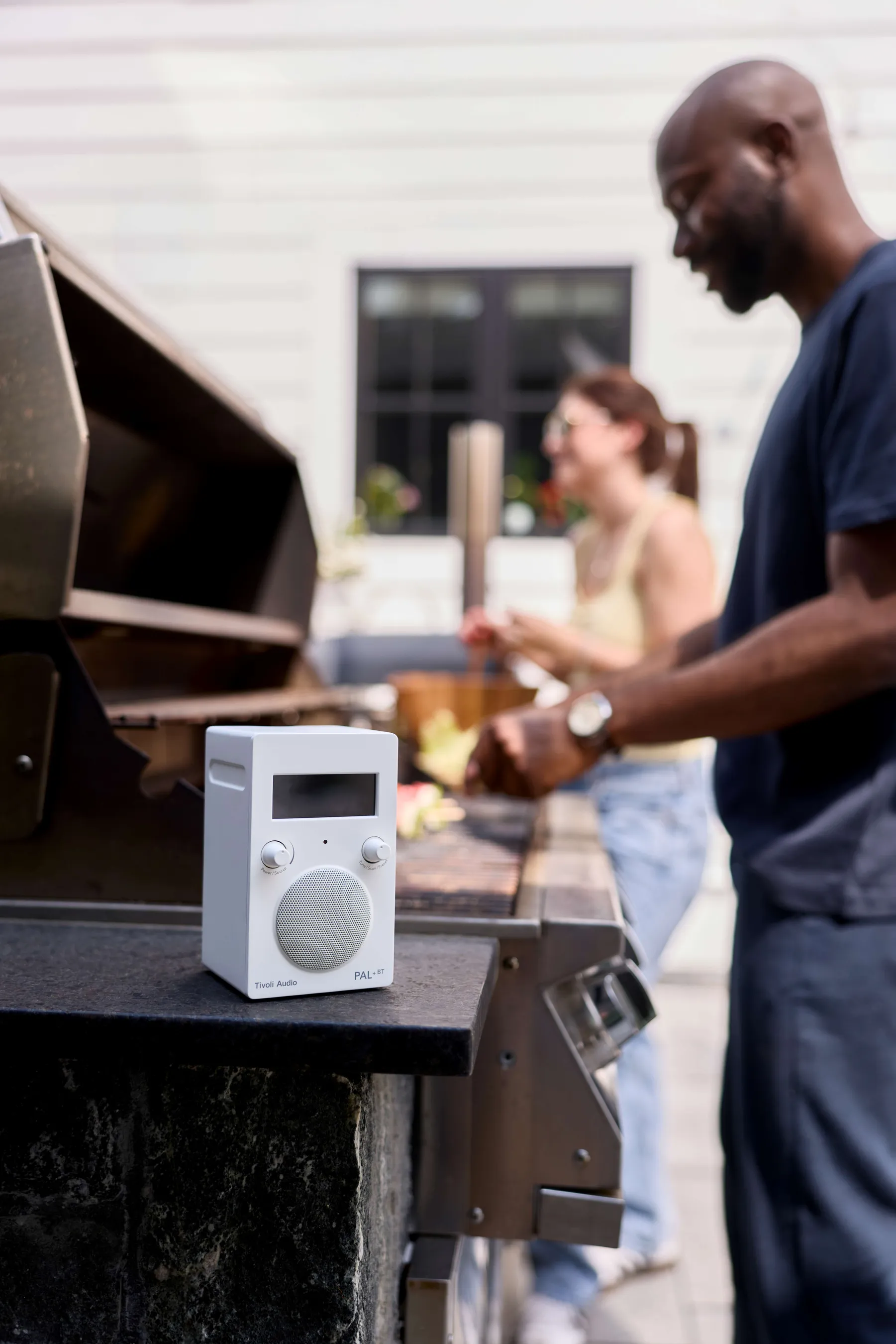 Un homme et une femme font un barbecue dehors. Une enceinte blanche est posée sur un comptoir noir.