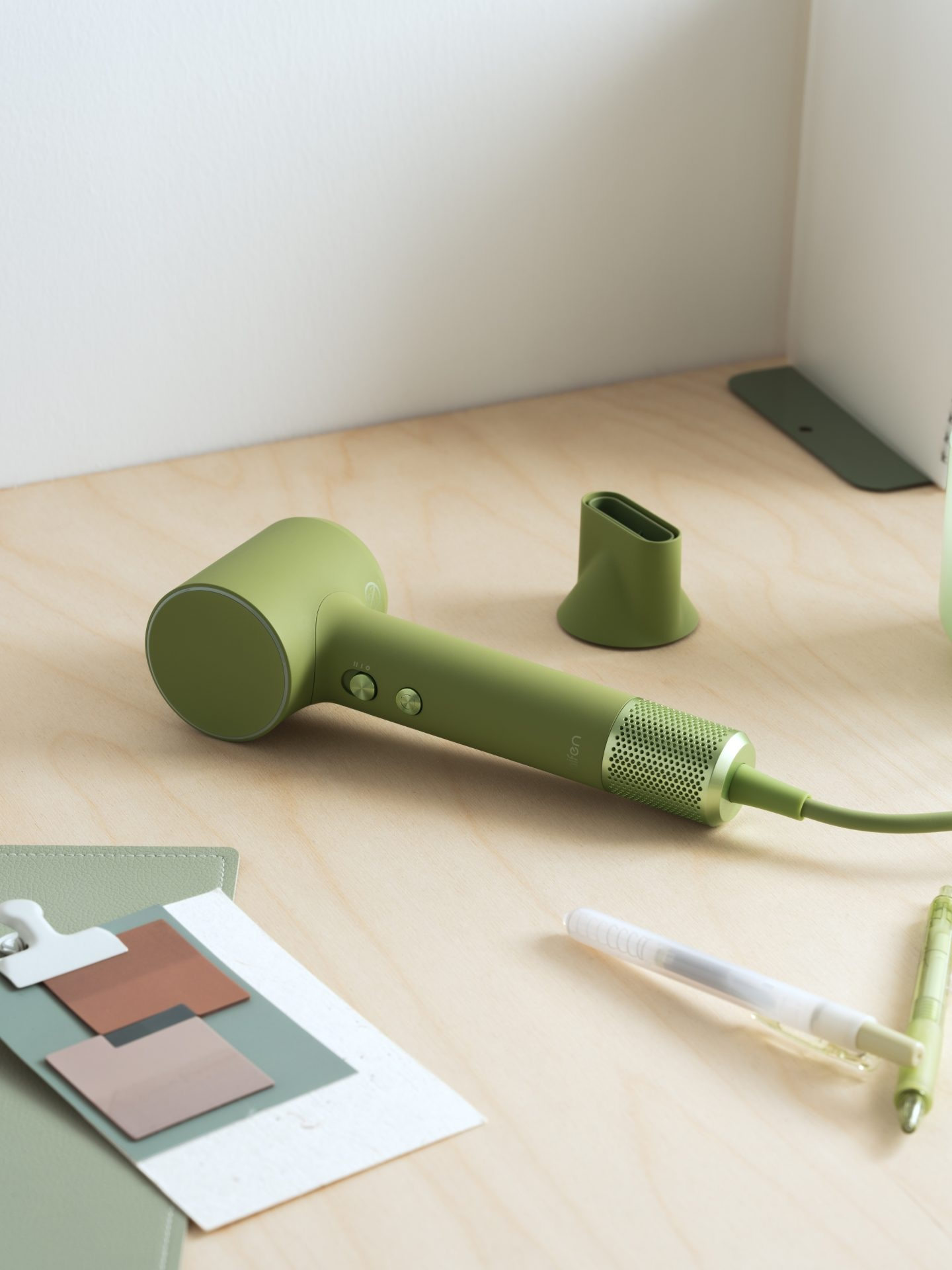 Zielona suszarka do włosów leży na drewnianym biurku obok próbek kolorów i długopisów.