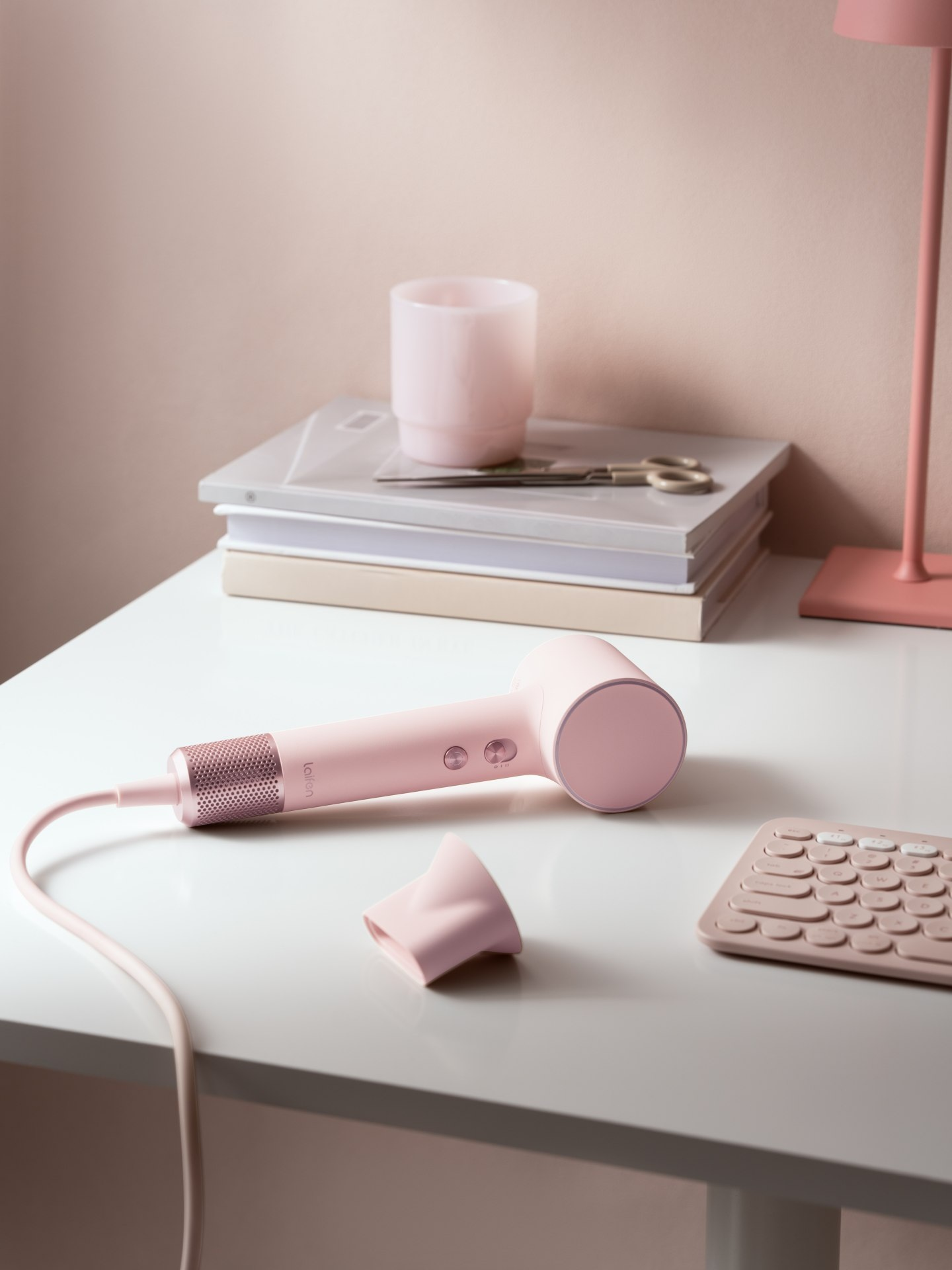 Różowa suszarka do włosów leży na biurku obok klawiatury i książek.