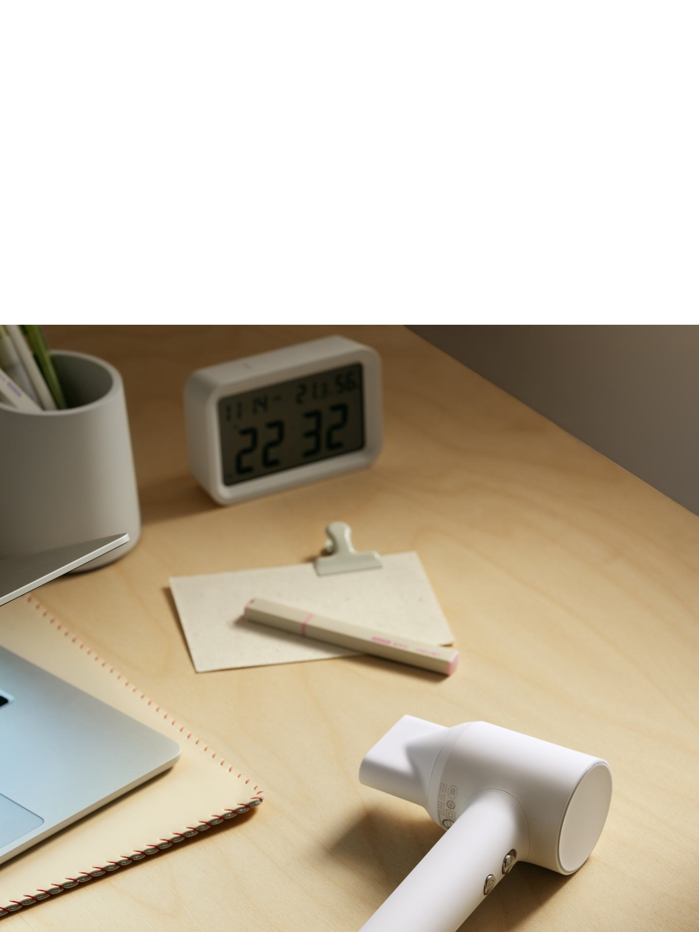 Biała suszarka do włosów leży na drewnianym biurku obok laptopa i zegara cyfrowego.