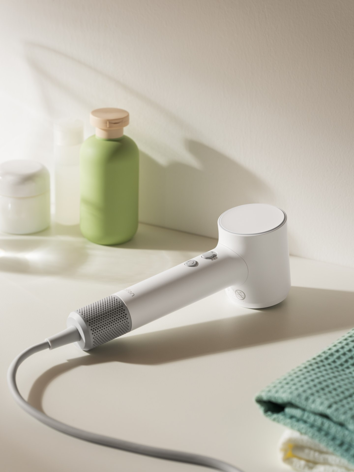 Biała suszarka do włosów leży na jasnej powierzchni obok butelek i ręczników.