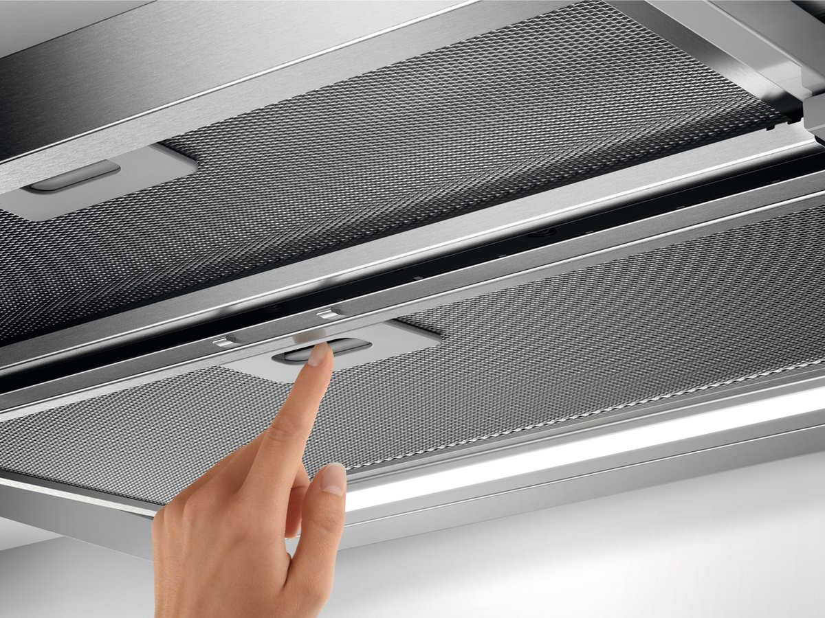 A finger presses a button on a metal mesh surface of a range hood.