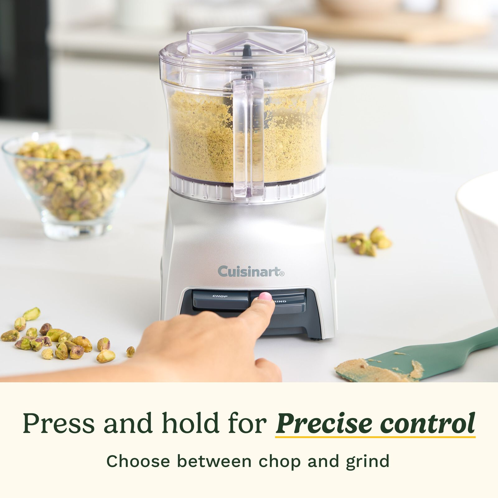 Eine Hand drückt die "Grind"-Taste eines Cuisinart-Zerkleinerers neben Pistazien.