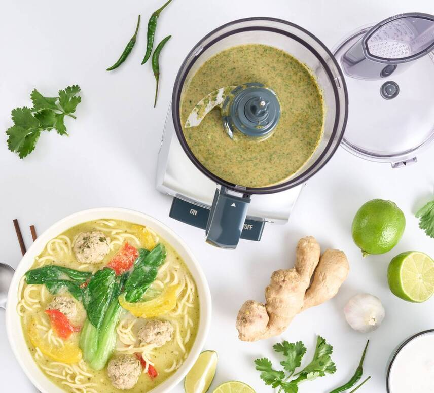Una ciotola di zuppa di noodle con polpette e verdure accanto a un robot da cucina.