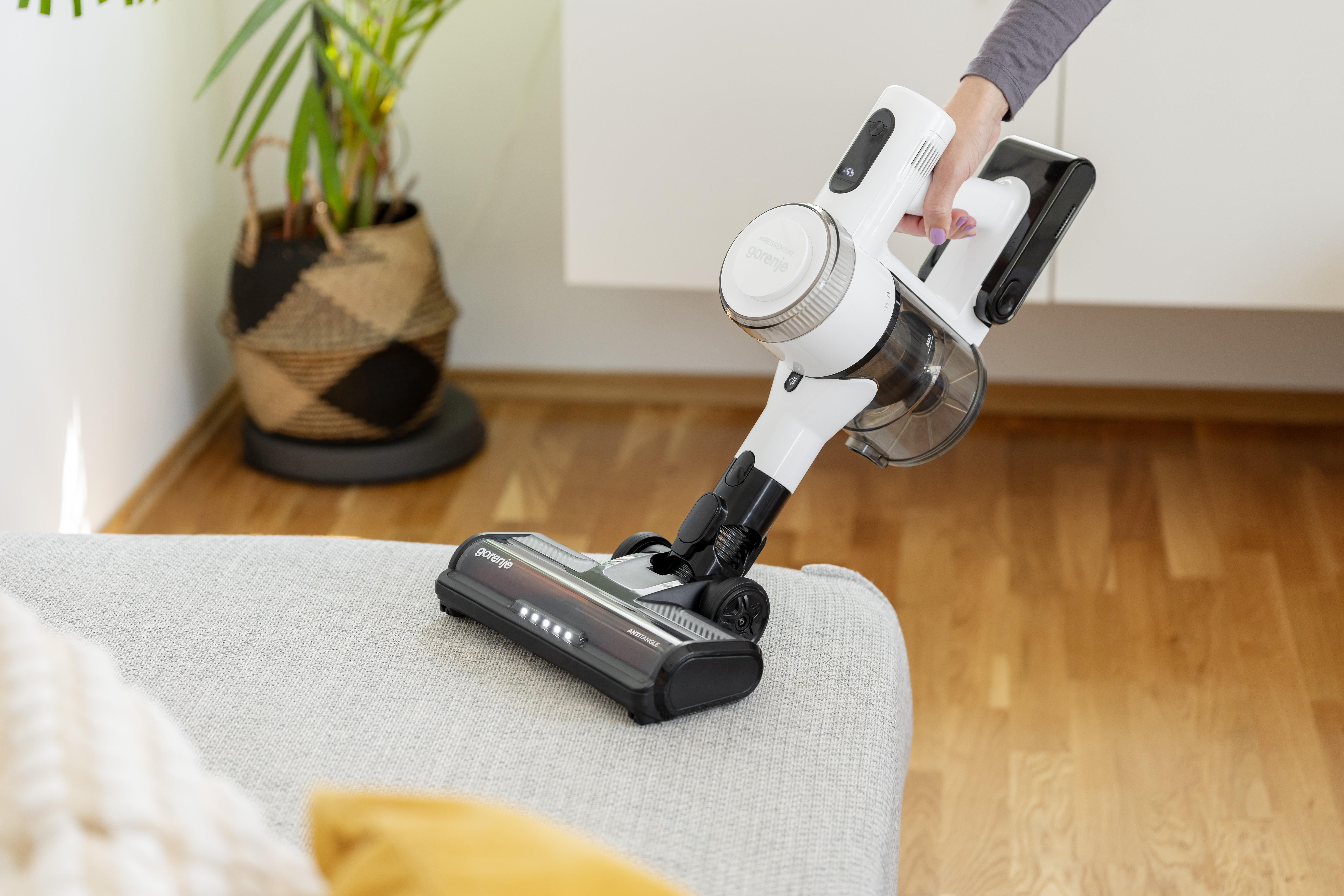 Eine Hand hält einen weißen Akku-Staubsauger über einem grauen Sofa.