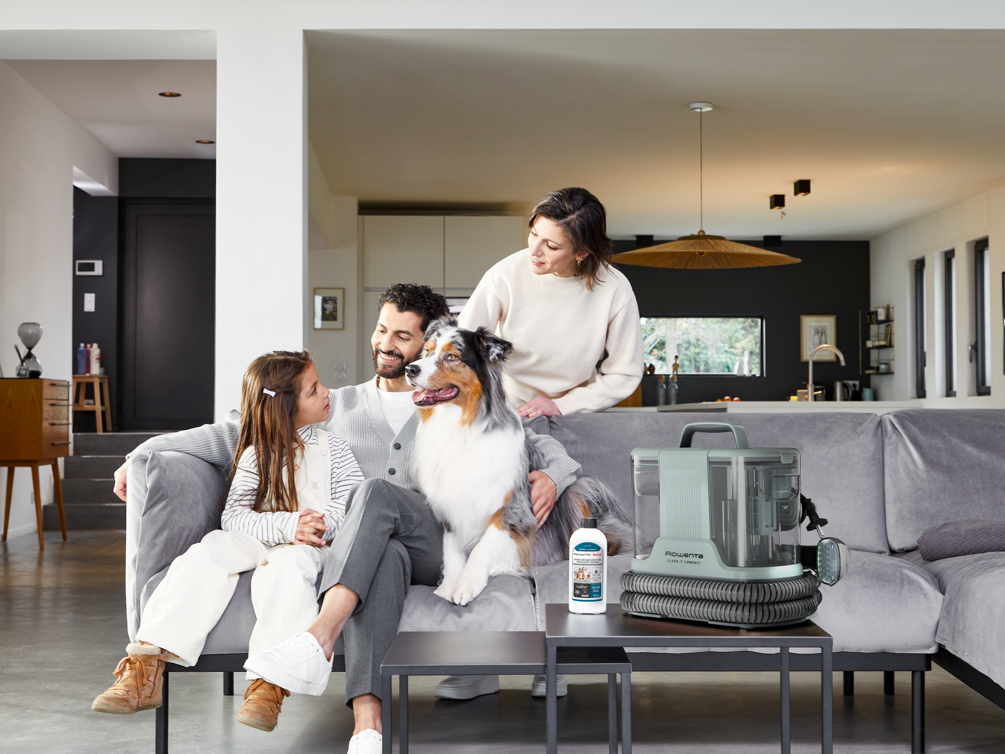 Eine Familie mit Hund sitzt auf einem Sofa, daneben ein Reinigungsgerät.