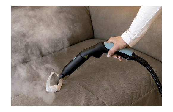 Una mano usa un limpiador a vapor en un sofá de tela marrón, liberando vapor.