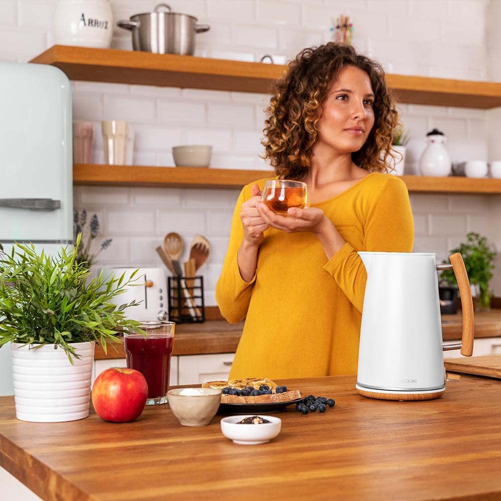 Mujer con blusa amarilla sosteniendo té en cocina con hervidor blanco, gofres, fruta.