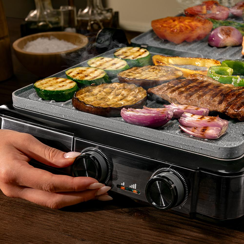 Una mano ajusta una parrilla eléctrica con carne y verduras cocinándose.