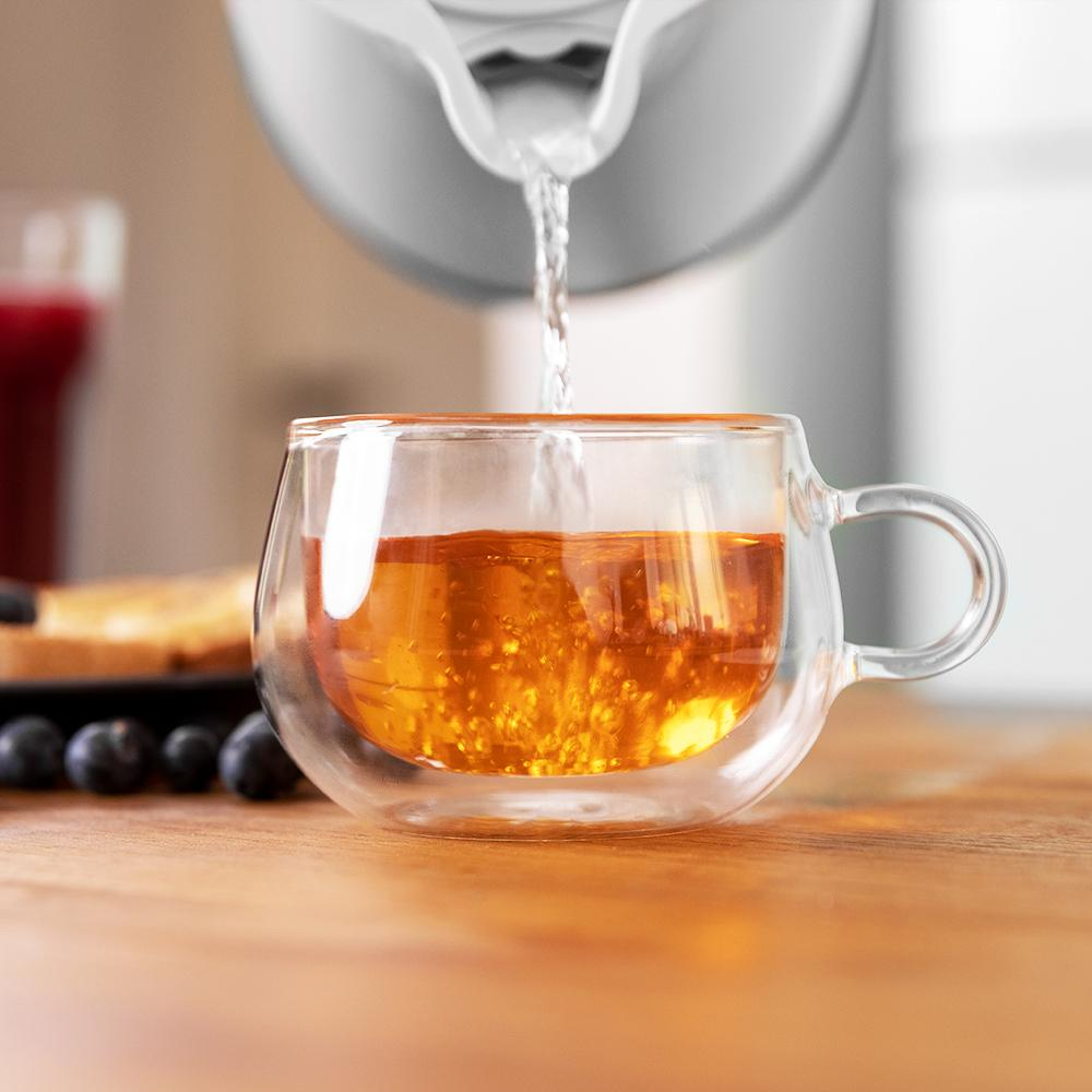 Se vierte agua caliente en una taza de vidrio, con tostadas y arándanos cerca.