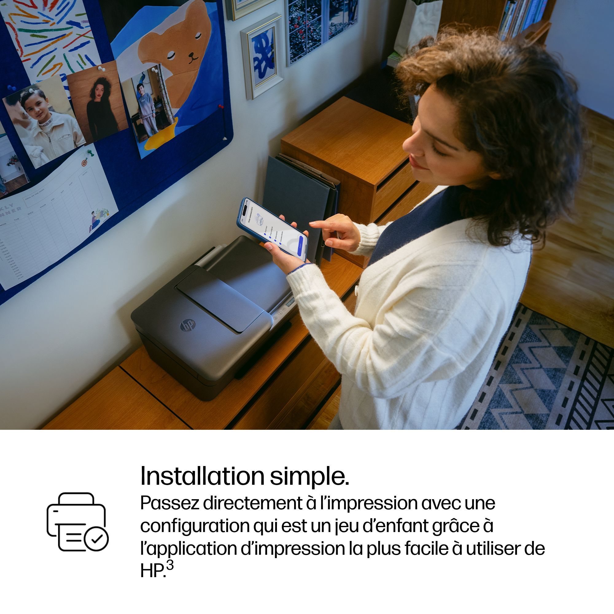 Une femme configure une imprimante avec son téléphone. Un agenda hebdomadaire est accroché au mur.