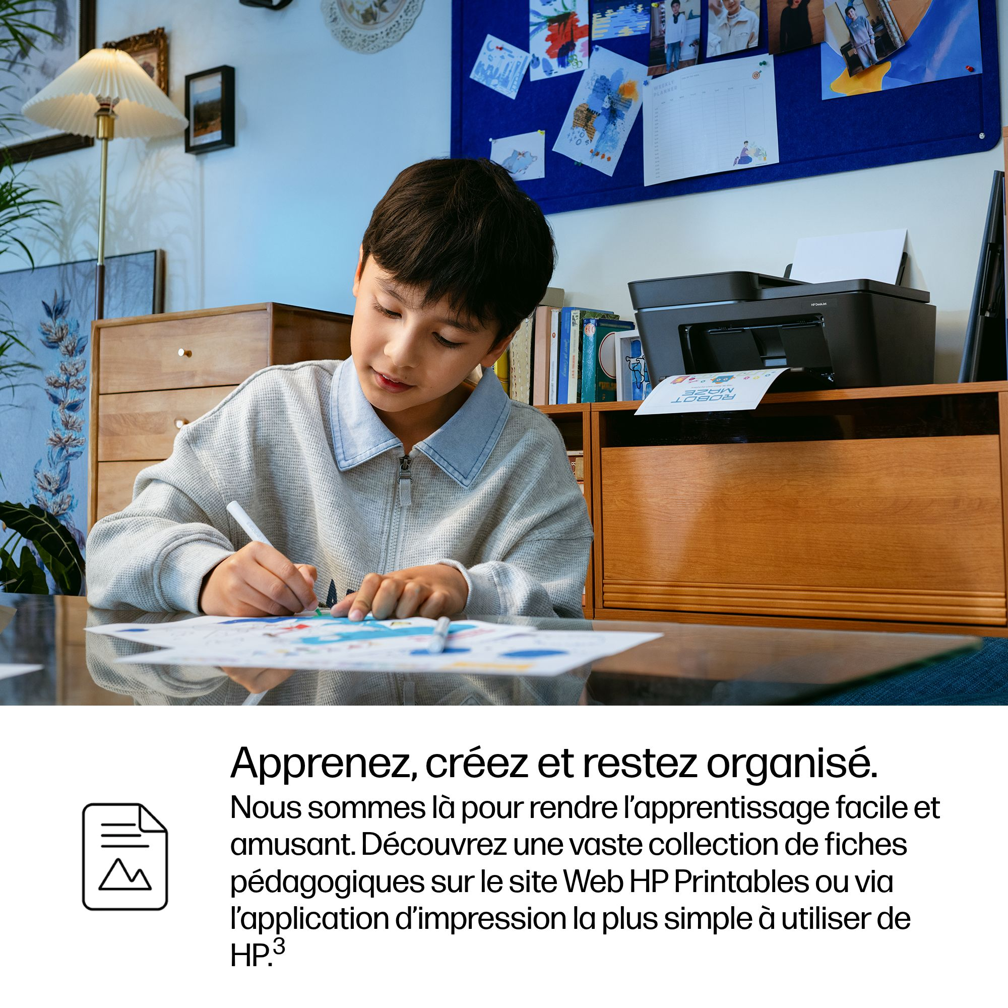 Un garçon dessine à un bureau, avec une imprimante et un tableau d'affichage à proximité.