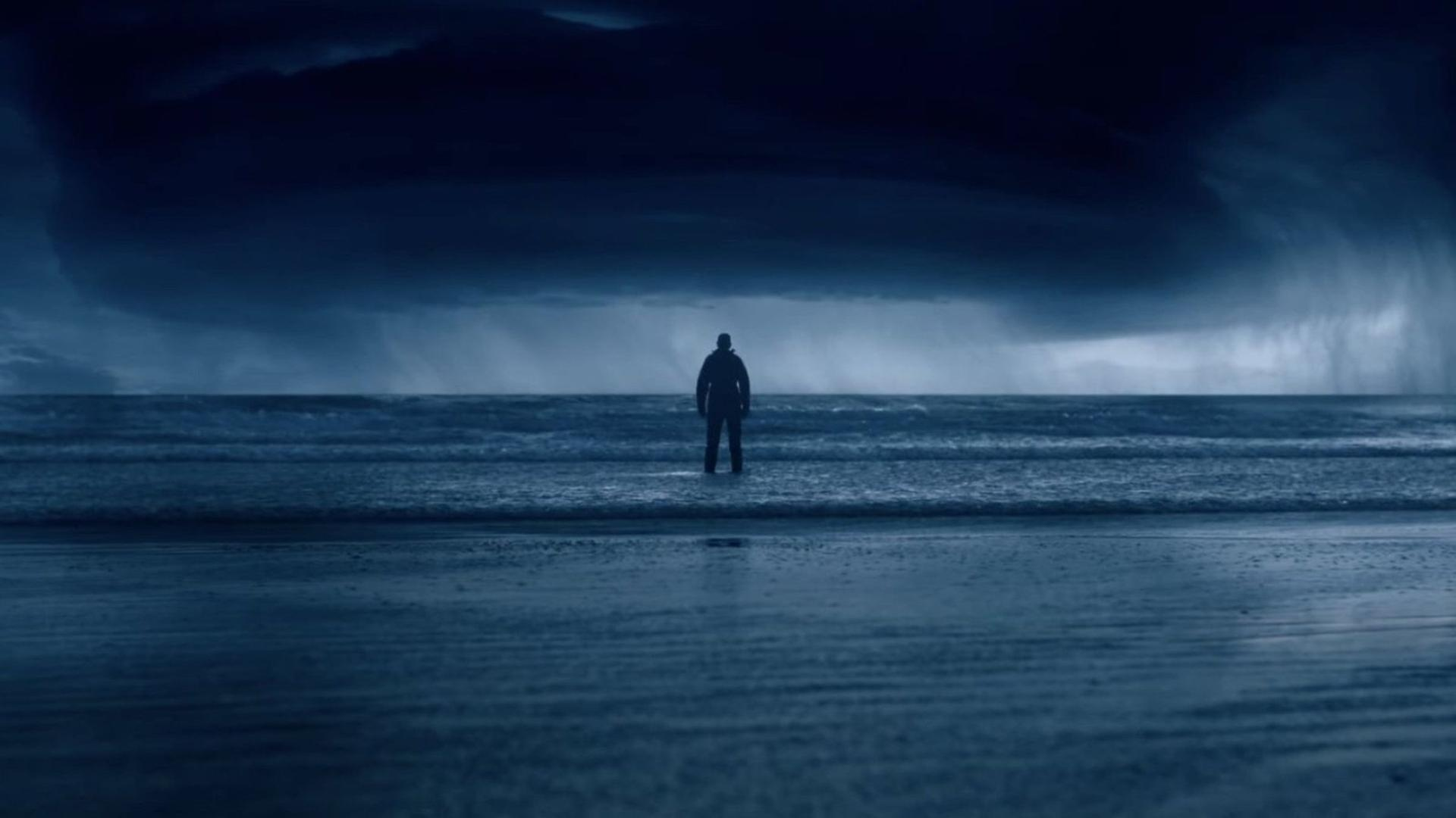 Eine Person steht im Meer unter einem stürmischen Himmel.