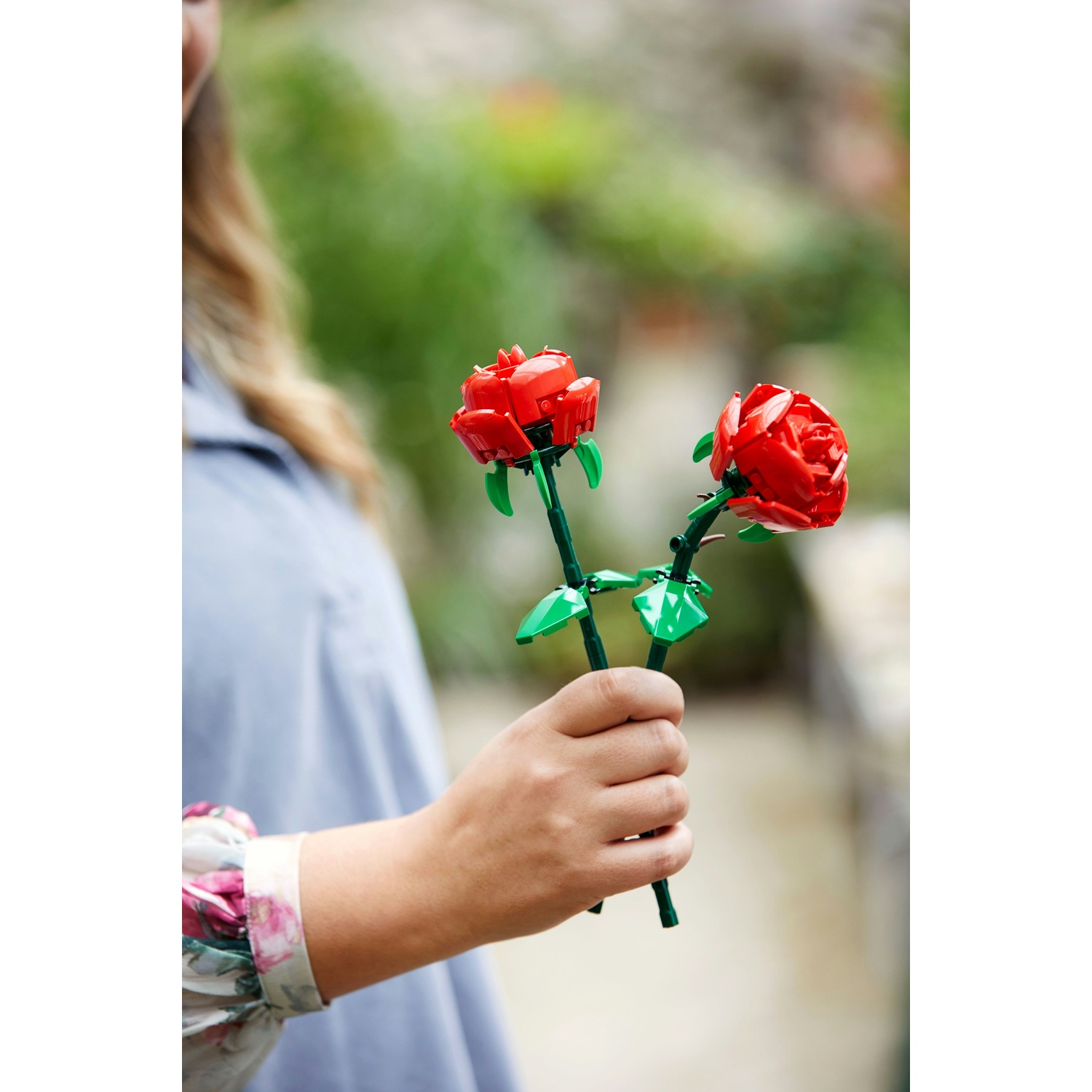 Persona tiene due rose rosse Lego con steli verdi. Sfondo sfocato.