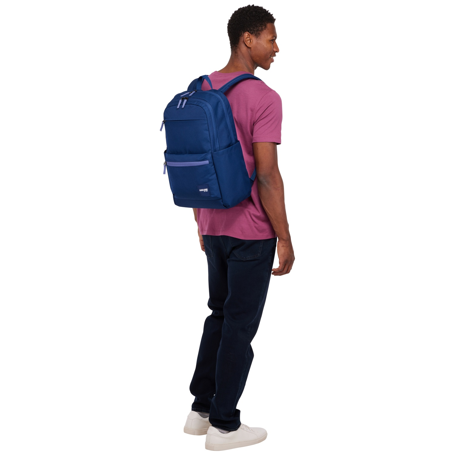 A man with dark hair wears a dark blue backpack and a pink shirt.
