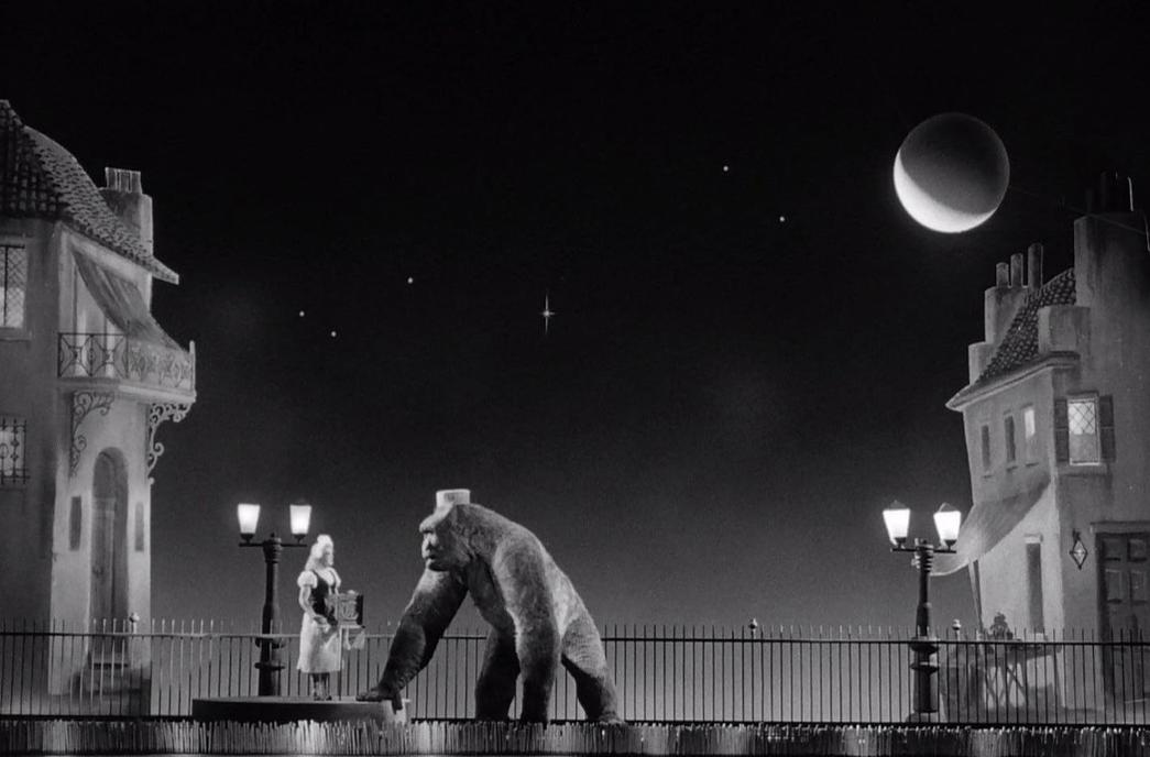 Ein Gorilla und eine Frau stehen nachts auf einer Promenade vor Häusern.
